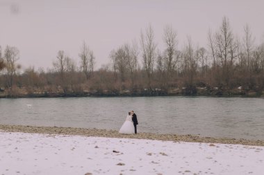 Bir gelin ve damat, karlı bir sahilde bir su kütlesinin yanında öpüşüyorlar. Sahne huzurlu ve romantik. Çiftin birlikte geçirdikleri özel anın tadını çıkarıyorlar.