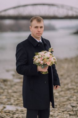 Siyah bir ceket giyen ve elinde bir buket çiçek tutan bir adam. Adam kayalık bir sahilde duruyor.