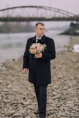 Plajda elinde bir buket çiçek tutan bir adam var. Sahne sakin ve huzurlu, adamın denize baktığı bir yer. Buketteki çiçekler pembe.