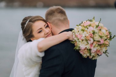 Bir gelin ve damat bir buket çiçek tutarken birbirlerini kucaklarlar. Gelin beyaz bir elbise ve kürk şort giyiyor ve damat da takım elbise giyiyor.