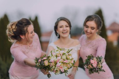 Üç kadın çiçek buketleri tutuyor ve gülümsüyor. Pembe giyinmişler ve özel bir olayı kutluyorlar.