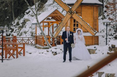 Gelin ve damat, beyaz bir elbise giymiş gelinle karda yürüyorlar. Sahne karlı bir alanda geçiyor, arka planda yel değirmeni var.