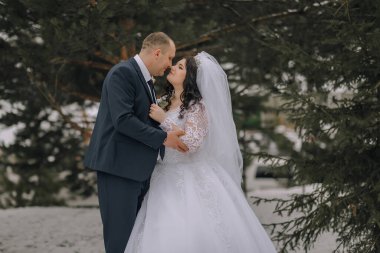 Bir gelin ve damat karda öpüşüyor. Gelin beyaz bir elbise giyiyor ve damat mavi bir takım giyiyor.