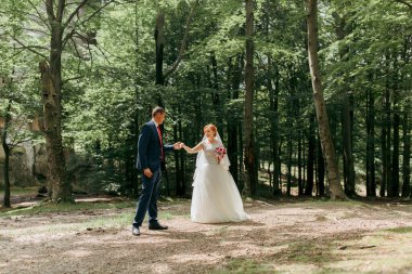 Bir gelin ve damat el ele bir ormanda yürüyorlar. Gelin beyaz bir elbise giyiyor ve damat mavi bir takım elbise giyiyor. Çift ağaçlarla çevrili ve atmosfer sakin.