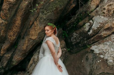 Beyaz elbiseli bir kadın kaya duvarının önünde duruyor. Yeşil bir saç bandı ve duvak takıyor.