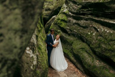 Bir çift mağarada fotoğraf çektirmek için poz veriyor. Adam mavi bir takım elbise giyiyor ve kadın beyaz bir elbise giyiyor. Sahne romantik ve samimi.
