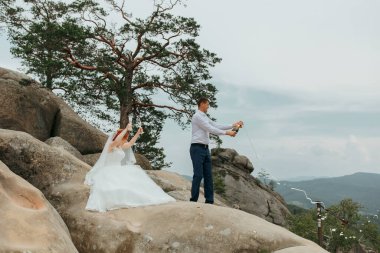 Bir gelin ve damat düğünlerini dağın tepesinde kutluyorlar. Gelin beyaz bir elbise giyiyor ve damat mavi bir takım elbise giyiyor. Ağaçlarla çevrili ve gökyüzü bulutlu.