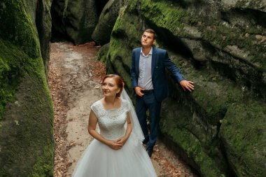 Gelin ve damat bir mağarada, gelin beyaz bir elbise ve damat mavi bir takım elbise giyiyor.