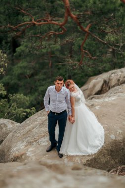 Gelin ve damat kayalık bir yamaçta duruyorlar, gelin beyaz bir elbise giyiyor ve damat mavi gömlek ve mavi pantolon giyiyor.