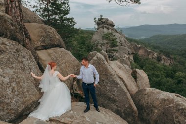 Gelin ve damat kayalık bir dağda duruyorlar, gelin beyaz bir elbise ve damat mavi bir gömlek giyiyor.