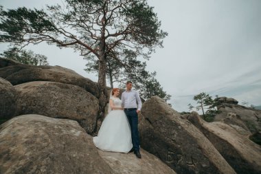 Bir çift kayalık bir yamaçta fotoğraf çektirmek için poz veriyor. Adam mavi bir gömlek giyiyor ve kadın beyaz bir elbise giyiyor. Macera ve romantizm kavramı