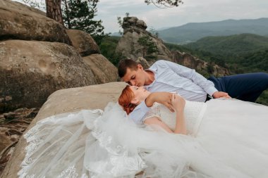 Bir gelin ve damat bir kayanın üzerinde uzanıyor, birbirlerini öpüyorlar. Gelin beyaz bir elbise giyiyor ve damat mavi bir gömlek giyiyor. Sahne huzurlu ve romantik.