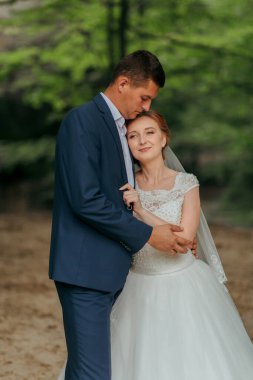 Bir adam ve bir kadın ormanda sarılıyorlar. Adam mavi bir takım giyiyor ve kadın beyaz bir elbise giyiyor.