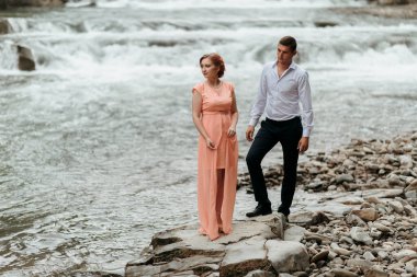 Nehrin kıyısındaki kayalık bir sahilde duran bir çift. Kadın pembe bir elbise giyiyor ve adam beyaz bir gömlek giyiyor.