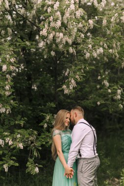 Arkalarında bir ağaç olan bir çift ormanda sarılıyor. Kadın yeşil bir elbise giyiyor ve adam beyaz gömlek ve pantolon askısı giyiyor.