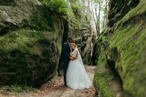 Bir gelin ve damat, yosun ve bitkilerle çevrili bir mağarada duruyor. Çift resmi giyinmiş, gelin beyaz bir elbise ve damat da takım elbise giymiş.