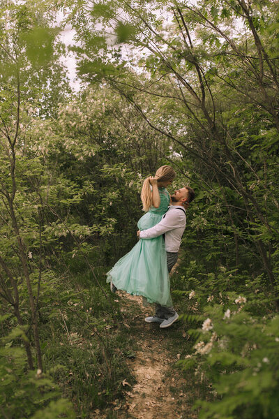 A couple is posing in a forest, with the woman holding the man up. Scene is romantic and playful