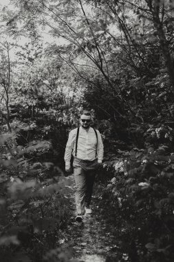 Beyaz gömlek ve pantolon askısı takmış bir adam ormanda yürüyor. Adamın etrafı ağaçlarla çevrili ve orman karanlık ve gölgeli olduğu için görüntüde tuhaf ve gizemli bir his var.