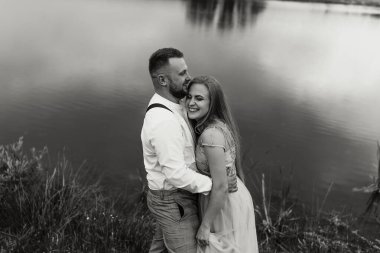 Bir çift göl kenarında, pantolon askısı takan adamla kucaklaşıyor. Kadın elbise giyiyor ve gülümsüyor.