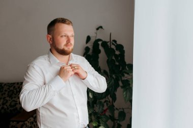 Beyaz gömlekli bir adam kravat takmaya hazırlanıyor. Pencereden dışarı bakıyor ve odada bir saksı bitkisi var.