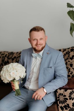 Mavi takım elbiseli ve beyaz gömlekli bir adam bir buket beyaz çiçek tutuyor. Gülümsüyor ve mutlu.