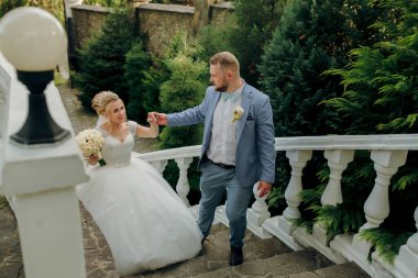 Bir gelin ve damat merdivenlerden iniyorlar. Gelin bir buket tutuyor ve damat elini tutuyor. Sahne mutlu ve romantik.