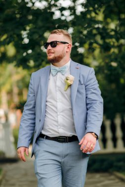 Mavi takım elbiseli ve beyaz gömlekli bir adam kaldırımdan aşağı yürüyor. Güneş gözlüğü ve papyon takıyor.