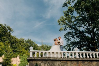 Gelin ve damat, gökyüzünün arka planında taş bir podyumda duruyorlar.