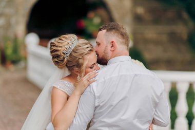 Bir gelin ve damat beyaz bir balkonda birbirlerine sarılıyorlar. Gelin taç takıyor ve damadın sakalı var.