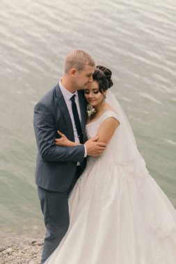 Bir gelin ve damat deniz kenarında bir plajda sarılıyorlar. Gelin beyaz bir elbise giyiyor ve damat bir takım elbise giyiyor. Sahne romantik ve samimi, aşkı yakalıyor.