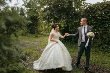 Bir gelin ve damat ormanda el ele tutuşup gülümsüyor. Gelin beyaz bir elbise giyiyor ve damat bir takım elbise giyiyor. Sahne mutlu ve romantik.