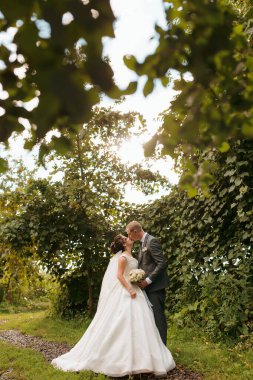 Bir gelin ve damat ormanda öpüşüyor. Gelin beyaz bir elbise giyiyor ve damat da takım elbise giyiyor.