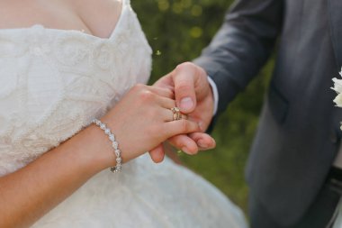 Bir gelin ve damat evleniyor ve gelin beyaz bir elbise giyiyor. Damat gelinin elini tutuyor ve ikisi de alyans takıyor.