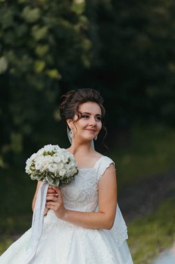 Elinde bir buket çiçek tutan beyaz elbiseli bir kadın. Gülümsüyor ve kameraya bakıyor.