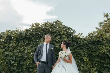 Gelin ve damat bir çitin önünde duruyorlar, gelin bir buket çiçek tutuyor.