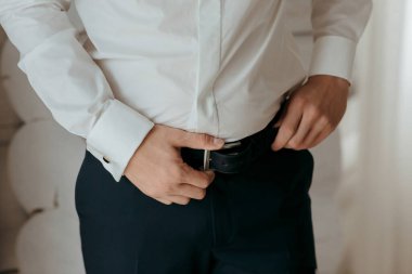 Beyaz gömlekli ve mavi pantolonlu bir adam kemerini ayarlıyor. Formalite ve detaylara dikkat etme kavramı. Çünkü adam kıyafetini düzgün bir şekilde giydiğinden emin olmak istiyor.