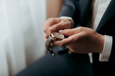 Bir adam takım elbise giyiyor ve elinde bir saat tutuyor. Saat altın ve deri bir bandı var.