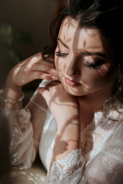 Uzun siyah saçlı ve beyaz elbiseli bir kadın güneşte oturuyor. Beyaz dantelli bir elbise ve bir bilezik giyiyor.