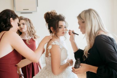 Bir gelin makyajını siyah elbiseli bir kadına yaptırıyor. Diğer kadınlar ona yardım ediyor.