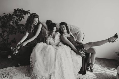 Üç kadın bir kanepede oturuyor, biri gelinlik giyiyor. Hepsi gülümsüyor ve birbirlerine eşlik etmekten zevk alıyor gibi görünüyorlar.