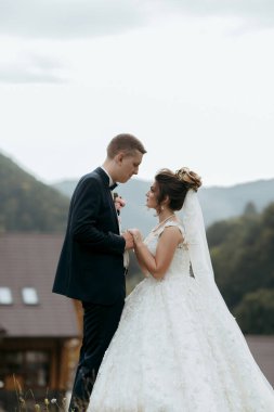 Gelin ve damat tepenin yamacında durur, gelin beyaz bir elbise giyer ve damat takım elbise giyer.