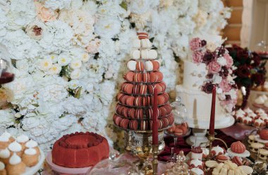 Arkasında beyaz bir duvar olan bir masa ve tepesinde de bir makaron kulesi. Makaronlar pembe ve beyazdır ve piramit şeklinde düzenlenmişlerdir. Masada başka tatlılar da var, pasta da dahil.