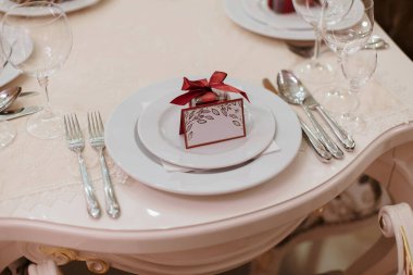 Beyaz masa örtüsü ve beyaz tabakta kırmızı bir fiyonk olan bir masa. Masa kırmızı bir kutu ve tabağında kırmızı bir kurdele olan özel bir durum için hazırlandı. Masada çatal bıçak takımı da var.
