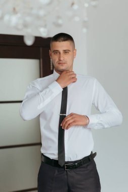 Beyaz gömlekli ve siyah kravatlı bir adam pencerenin önünde duruyor. Kravatını düzeltiyor ve kameraya bakıyor. Adam takım elbise giydiği için resmiyet ve profesyonellik kavramı.