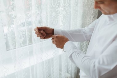 Beyaz perdeli bir adam pencerenin önünde giyiniyor. Gömleğini ve kravatını düzeltiyor.