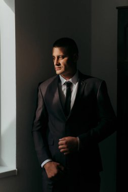 Takım elbiseli ve kravatlı bir adam pencerenin önünde duruyor. Kravat ve saat takıyor.