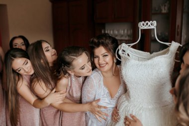 Bir grup kadın gelinliğe sarılıyor. Gelin gülümsüyor ve diğer kadınlar elbiseye bakıyor.