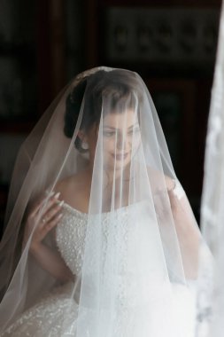 Bir kadın peçe takıyor ve gülümsüyor. Beyaz bir elbise giyiyor ve kollarını boynuna dolamış.