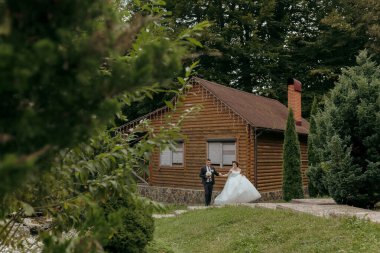 Bir gelin ve damat ahşap bir evin önünde duruyor. Gelin beyaz bir elbise giyiyor ve damat bir takım elbise giyiyor. Sahne romantik ve samimi.