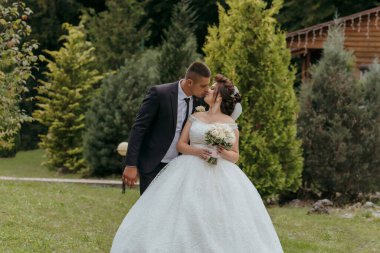 Parkta bir gelin ve damat öpüşüyor. Gelin beyaz bir elbise giyiyor ve damat da takım elbise giyiyor.